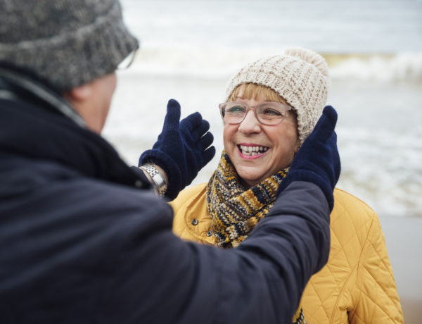 Senior couple walking on a beach together in winter. They are in the North East of England and the husband is adjusting his wife's hat for her.