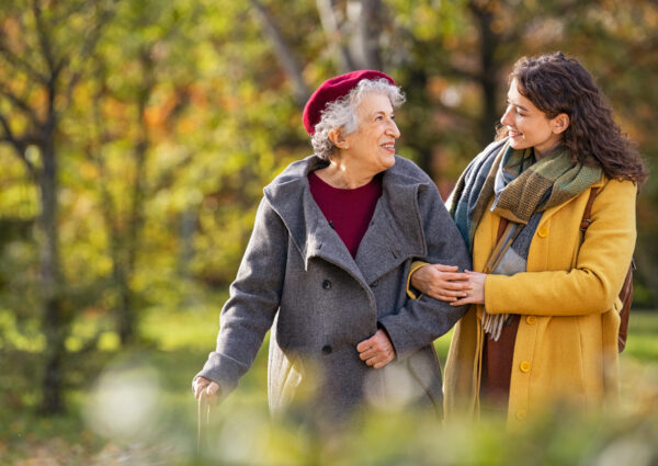 Young woman in park wearing winter clothing walking with old grandmother. Happy grandma wearing coat walking with lovely girl outdoor with copy space. Smiling lovely caregiver and senior lady walking in park during autumn and looking at each other.
