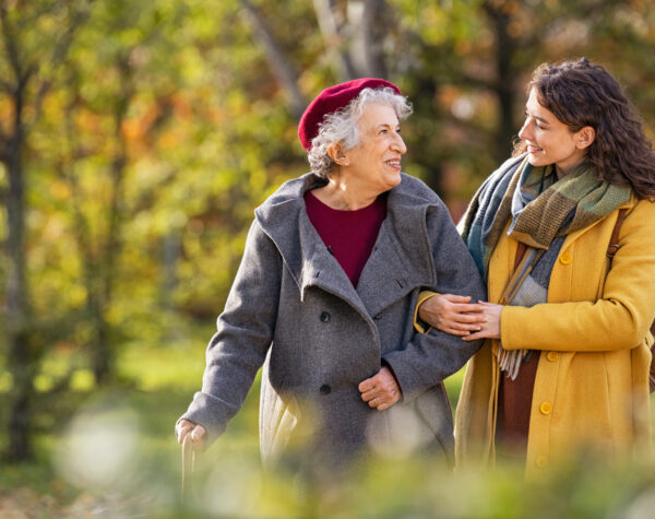 Young woman in park wearing winter clothing walking with old grandmother. Happy grandma wearing coat walking with lovely girl outdoor with copy space. Smiling lovely caregiver and senior lady walking in park during autumn and looking at each other.