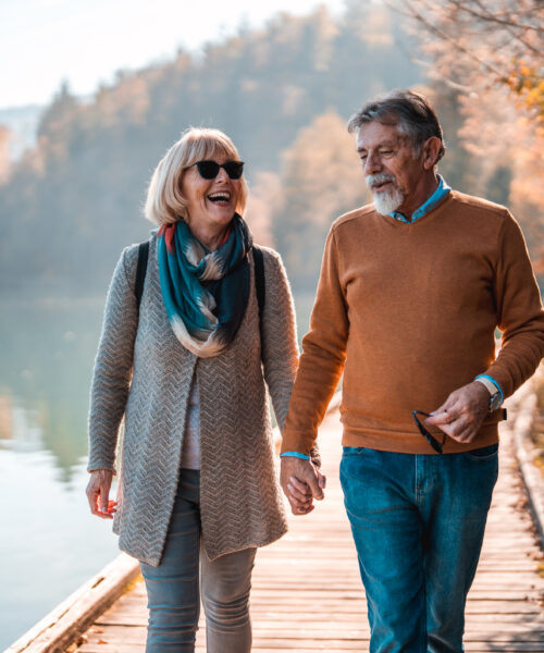 Female and Male Caucasian Senior Citizens Walking Hand in Hand Caucasian senior woman holding hands with her male companion taking a walk on a lakefront in European Alps. 3/4 length image, laughing while discussing something. Reflections in the water in the background.